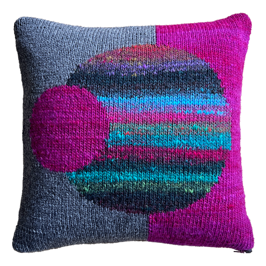 The hand-knit side of this pillow features a split background of charcoal gray (left) and magenta (right), punctuated by a large multicolored, striped sphere in the center and a smaller magenta sphere, off-centered.