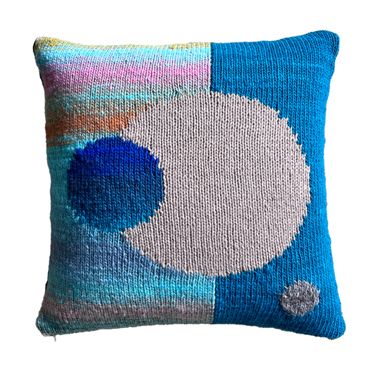 The hand-knit side of this pillow features a split background of stripes in shades of blue & pink (left) and turquoise (right), punctuated by a large neutral, driftwood sphere in the center and a smaller blue sphere, off-centered.