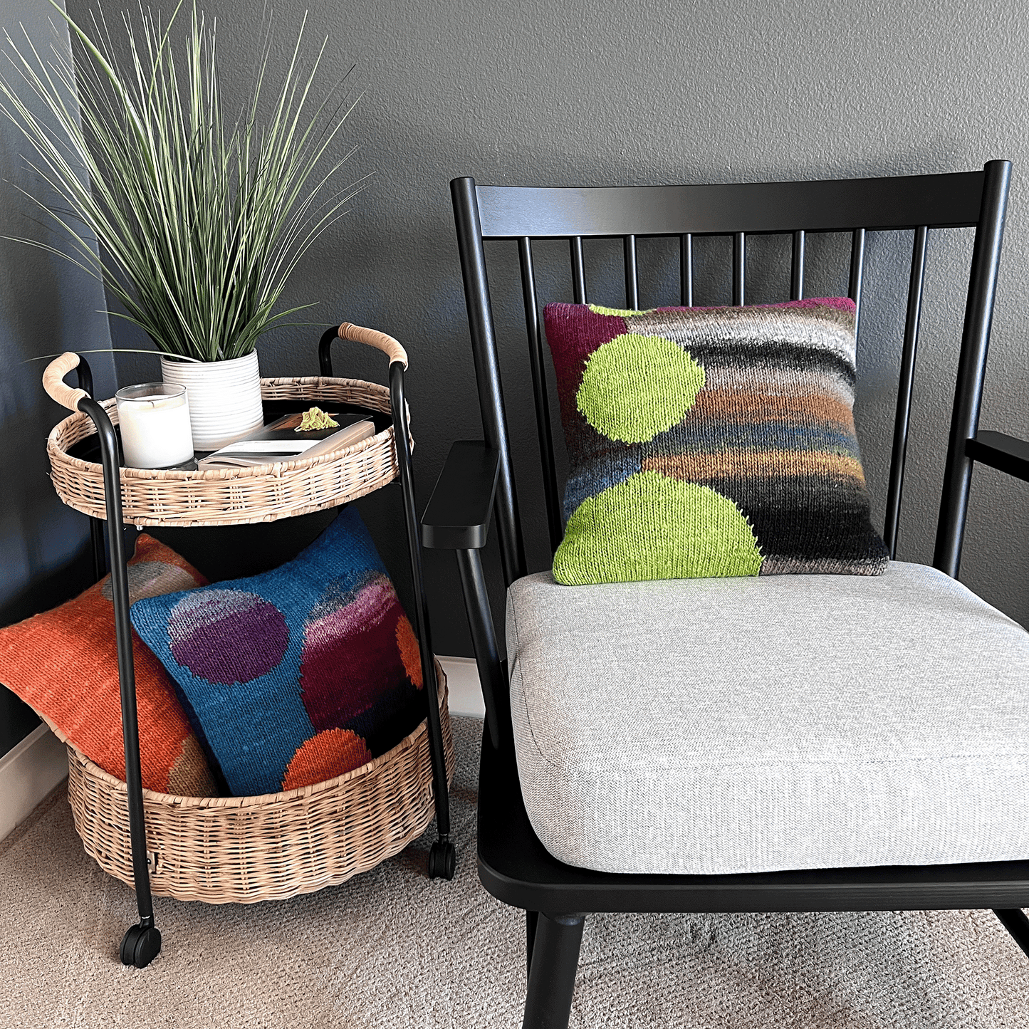 Three decorative throw pillows, hand-knit by Susan in 100% wool, display bold graphics in vibrant hues, shown against a black shaker chair.