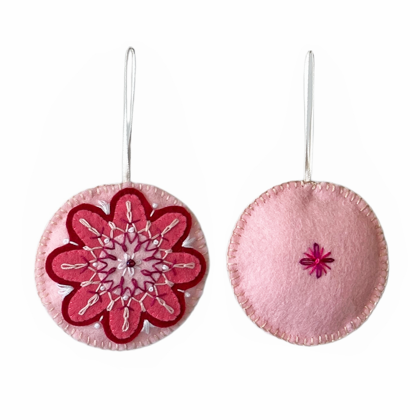 This Folk Flower ornament includes threes shades of pink felt, layered to create a floral shape on the front of a stuffed light pink circle. Both sides are embroidered with palest pink, white, and magenta threads. A single glass beads appears on the front.