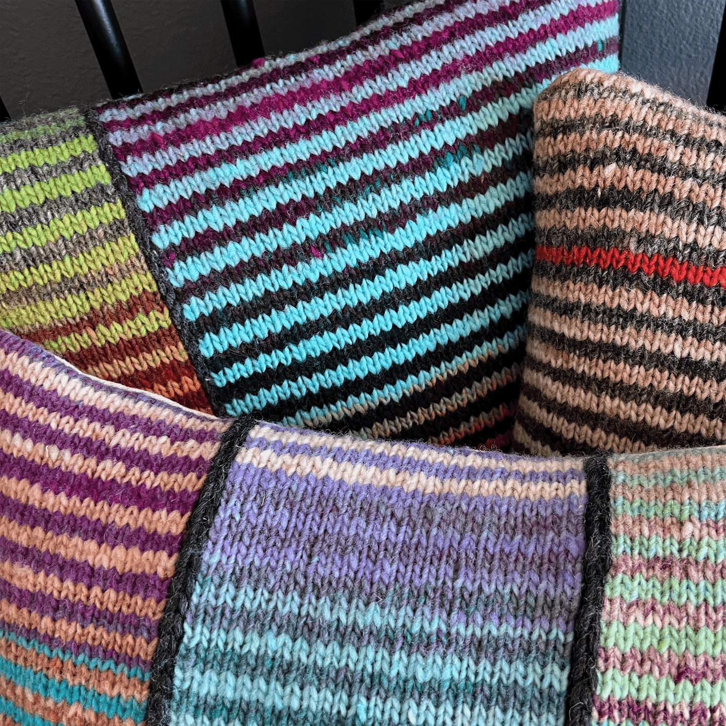 Three decorative throw pillows, hand-knit by Susan in 100% wool, display bold stripes in vibrant hues against a black shaker-style chair.