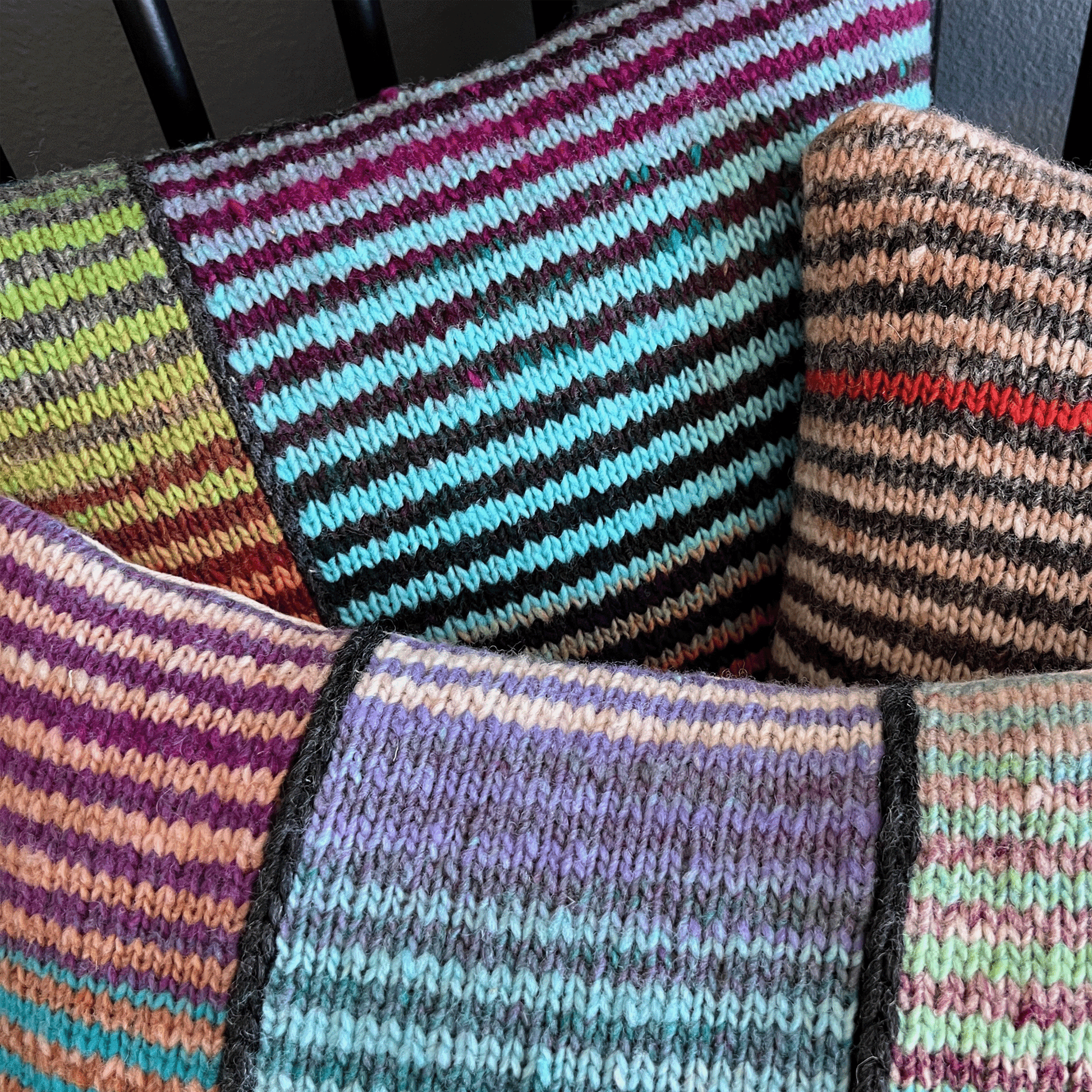 Three decorative throw pillows, hand-knit by Susan in 100% wool, display bold stripes in vibrant hues against a black shaker-style chair.