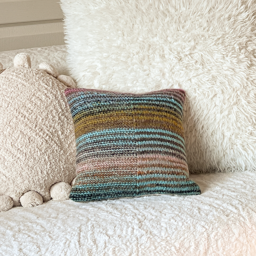 This 100% wool, hand-knit decorative throw pillow with pastel stripes pops against an off-white, shabby chic, boho-style couch.