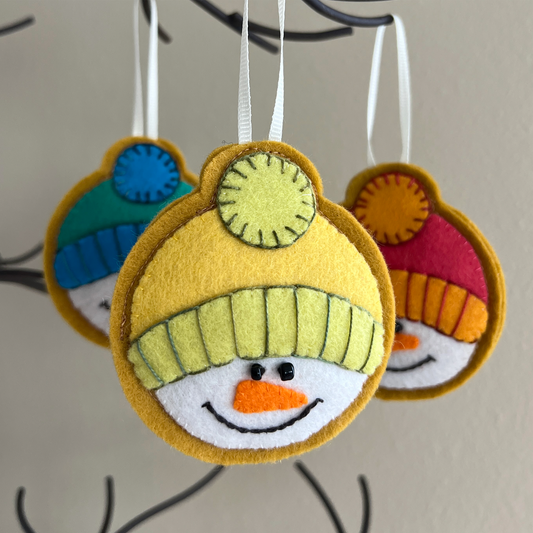 This wool felt ornament features the smiling face of a snowman with a cute carrot nose and twinkly black bead eyes. He's wearing a winter hat in two shades of yellow. The brim and pouf are embellished with a contrasting thread.
