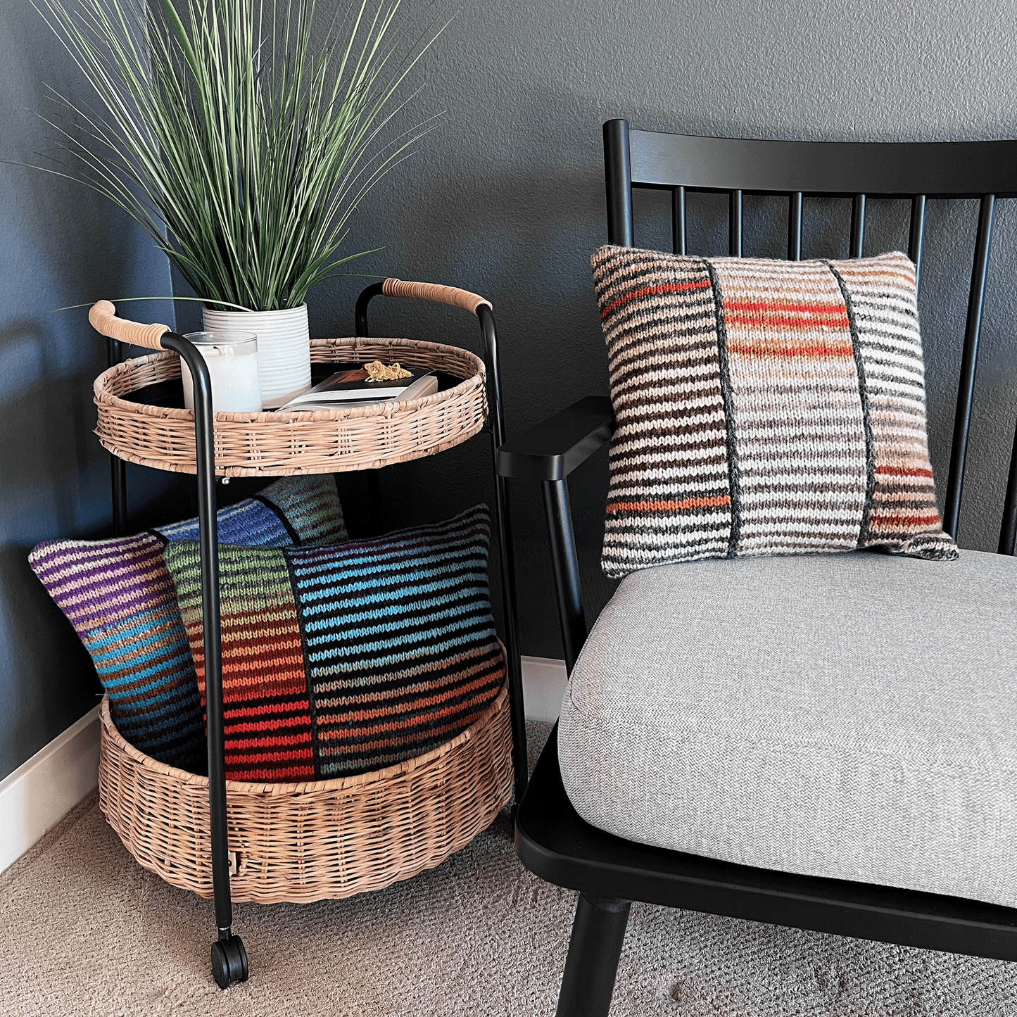 Three decorative throw pillows, hand-knit by Susan in 100% wool, display bold stripes in vibrant hues against a black shaker-style chair.