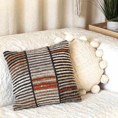 This 100% wool, hand-knit decorative throw pillow with variegated stripes pops against an off-white, shabby chic, boho-style couch.