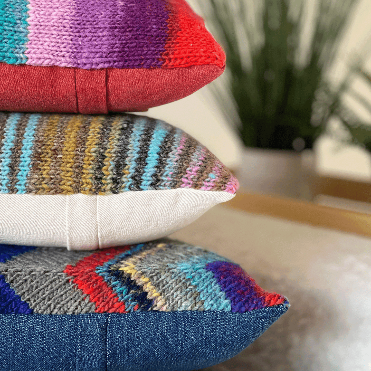 A stack of three decorative throw pillows, hand-knit by Susan in 100% wool, display bold stripes and geometric chevrons in vibrant hues.