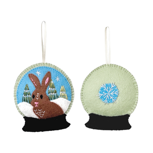 This Winter Animal ornament features an adorably plump brown bunny made of 100% wool felt, sitting in a snow-covered forest within a pale green snow globe. On the back is a hand-embroidered snowflake.