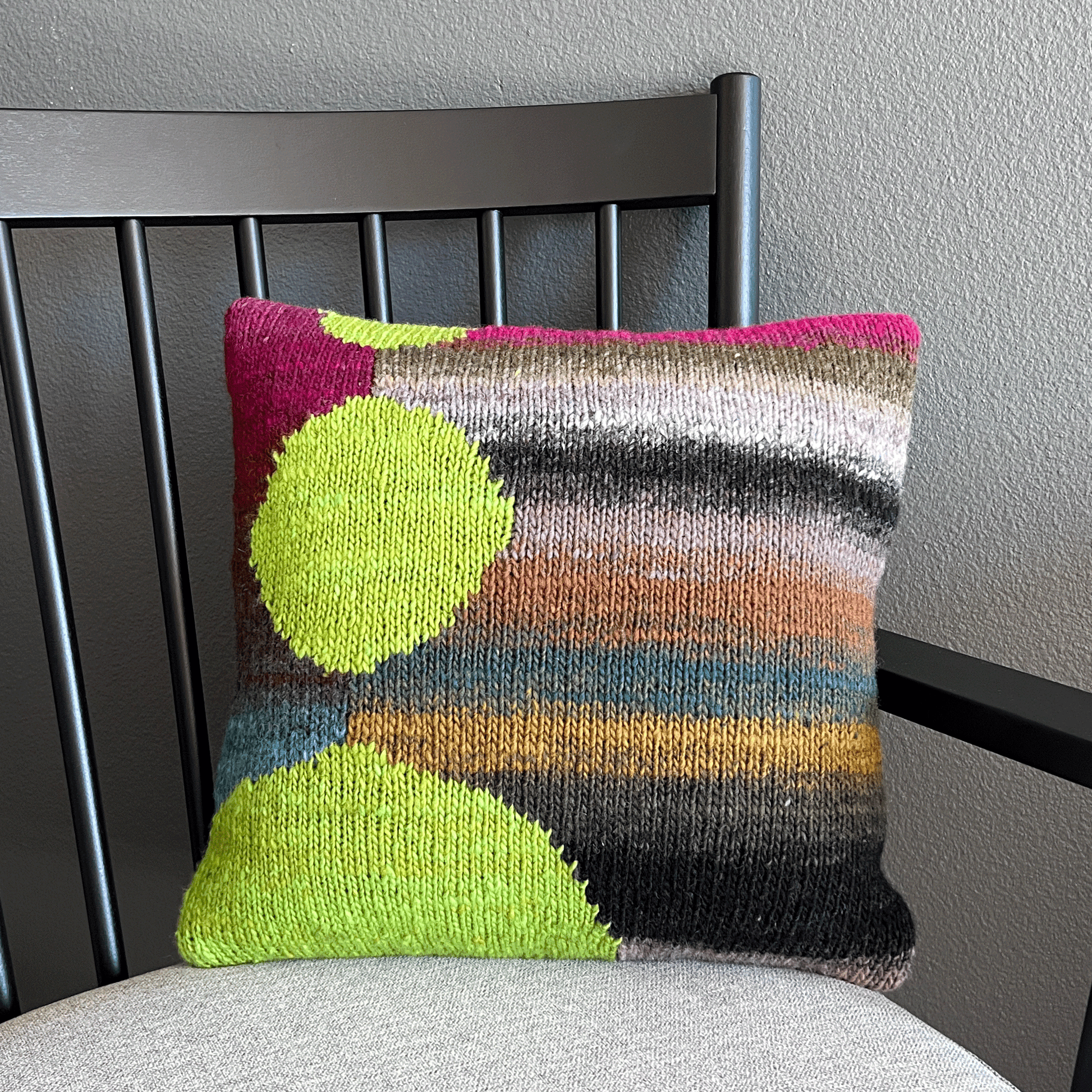 Gracing a black, shaker-style chair against a dark wall, this intarsia pillow's bright lime spheres really "pop" against its background of variegated stripes in rich earthy hues.